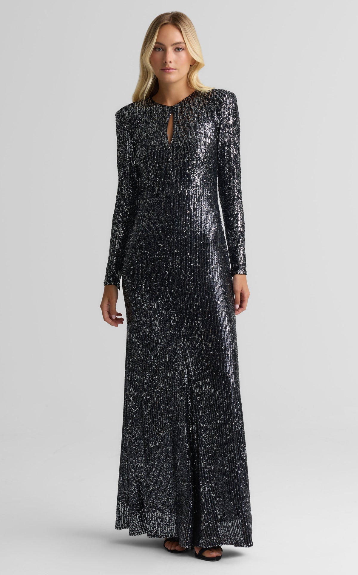 A woman modeling a floor-length black sequined gown with long sleeves, a round neckline with a small keyhole cutout, and a subtle front slit, photographed against a light gray studio background.