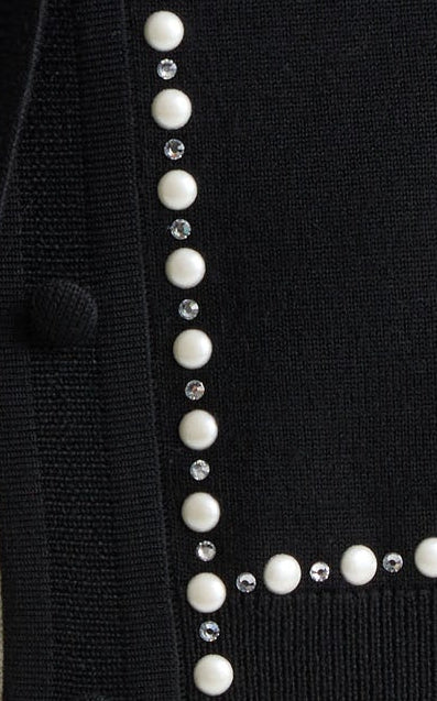 A blonde woman wearing a black knit cardigan with white pearl-like trim along the collar, front opening, cuffs, and hem, paired with cream pants, against a light gray studio background.