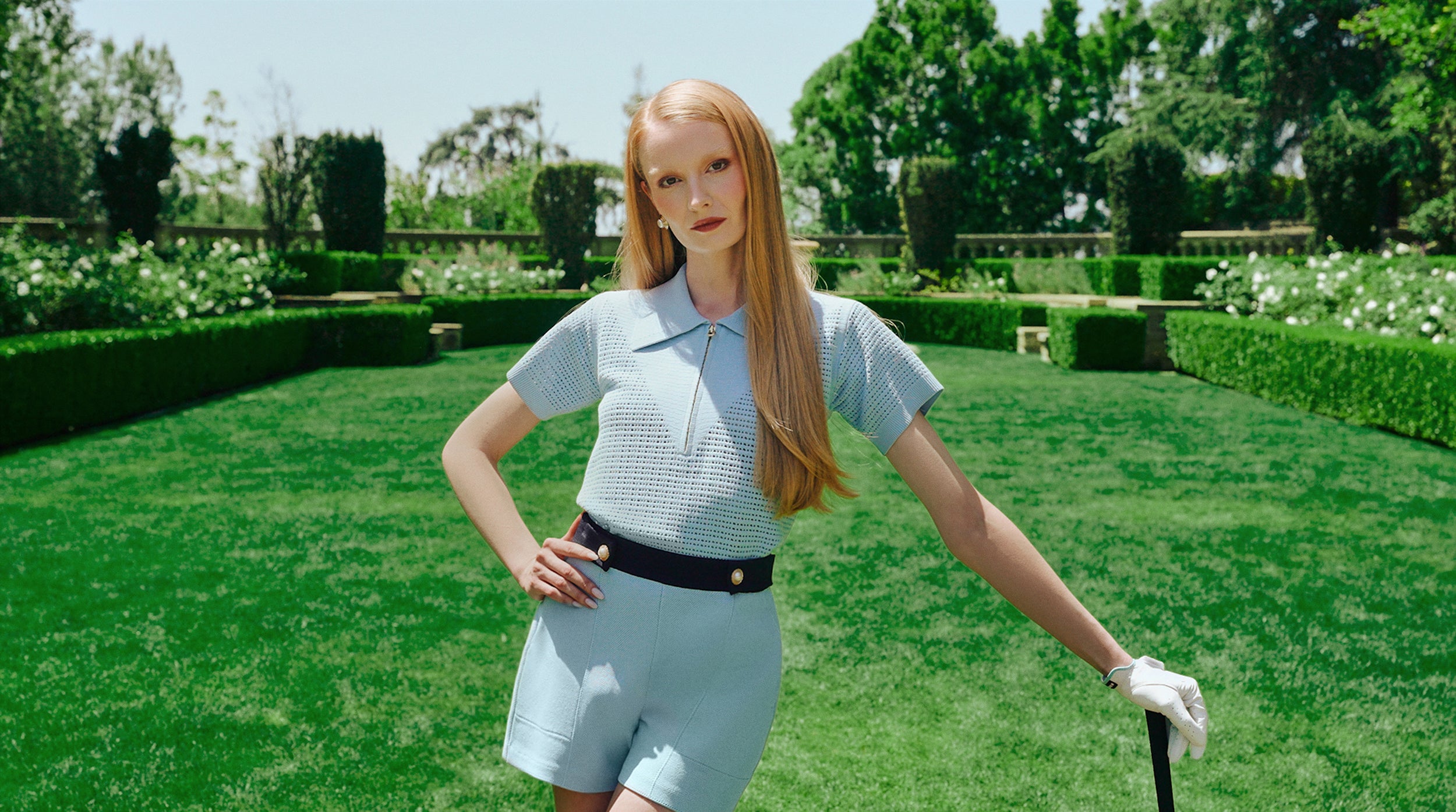 Woman in a light blue luxury golf outfit standing on a grassy lawn with manicured hedges and trees in the background.